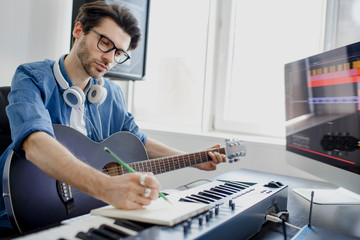 Male music arranger composing song on midi piano and audio equipment in digital recording studio. Man plays guitar and produce electronic soundtrack or track in project at home. - obrazy, fototapety, plakaty