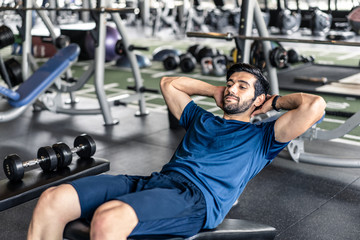 Fototapeta na wymiar Young fit handsome smiling Caucasian man exercising by doing a sit-up on bench in gym or fitness club. Male athlete has good body shape with six pack. Sport recreation and fitness for health concept.