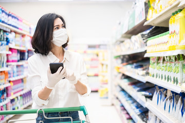 Asian woman wearing face mask and rubber glove push shopping cart in suppermarket departmentstore. Girl hold smartphone choose & look grocery things to buy during coronavirus crisis, covid19 outbreak.
