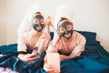 Two female models make face mask and using smartphone or phone for selfie and communication in social networks. Two young women in towels and pajamas have a fun spa party together in home.