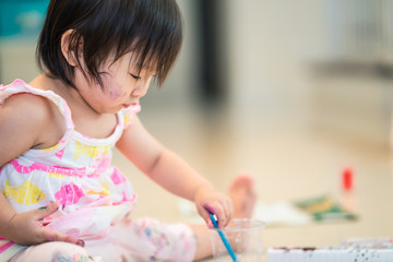 Asian cute baby girl intend to paint watercolor on paper in painting class at home. She holding paintbrush in the hand and painting animal from imagination. Happy and fun creative activity concept.