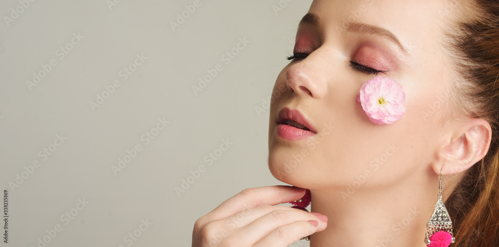 Wall mural Girl with a gentle make-up and a flower on her face. Delicate skin, skin care in the spring. Spoil the model, spring, flowering, beauty and personal care. A close-up of a pink tender flower on face.