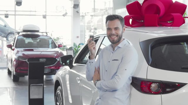 Handsome male driver holding car keys to his new auto at the dealership. Attractive male driver leaning on his new auto, showing car keys to the camera proudly. Car owner concept