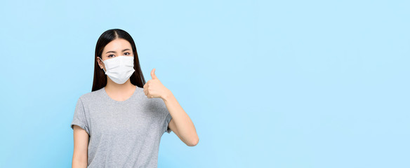 Young Asian woman wearing medical face mask and giving thumbs up isolated on light blue banner...