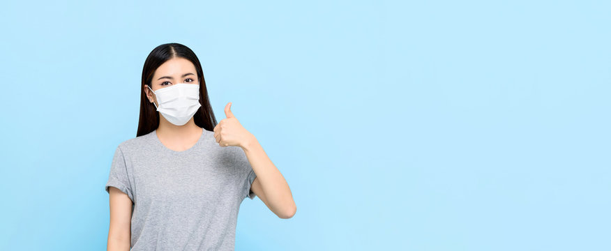 Young Asian Woman Wearing Medical Face Mask And Giving Thumbs Up Isolated On Light Blue Banner Background With Copy Space