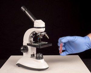 Microscope and hand with surgical glove holding vial with red solution. Black background with space for text