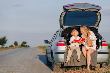 pretty blond hair woman with little blond son at sunset relaxing behind the car and eating watermelon. summer, travel, nature and fresh air in the countryside.