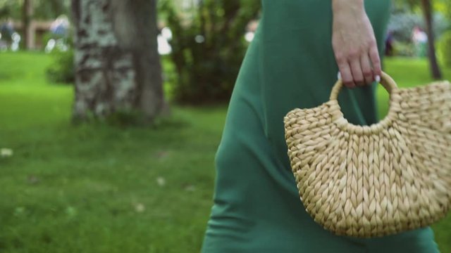 Close up of young woman with handmade stylish straw handbag walks in the summer park. Eco bag.