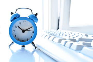 Computer technology and business concept white wallpaper. Blue classic alalrm clock showing morning time and white PC keyboard on blurred background of half open window shooting in high key 
