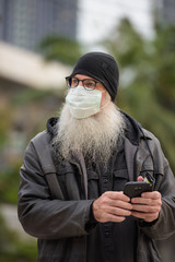 Mature bearded hipster man with mask thinking while using phone outdoors