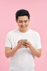 Waist-up portrait of joyful Vietnamese man leaning on gray wall while browsing Internet on smartphone, studio shot