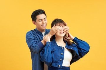 Guess who. Playful young guy standing behind girlfriend and closing her eyes, yellow studio background with copy space