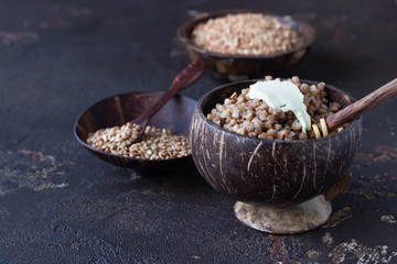 Coconut bowl with tasty boiled buckwheat and uncooked organic buckwheat grains in coconut bowls, dark brown concrete background. Gluten free, healthy diet.