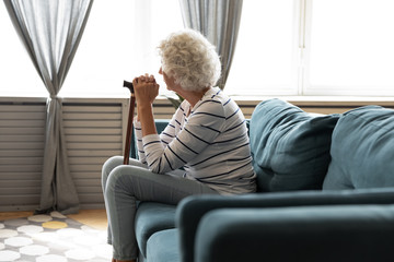 Rear view pensive older woman holding wooden cane, sitting on couch in living room alone, looking in window, thoughtful mature senior female using walking stick during rehabilitation