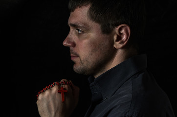 Portrait of a young man who praying with prayer beads with a crucifix on a cross on a black background
