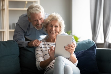 Happy older couple using computer tablet together at home, excited mature man and woman looking at mobile device screen, shopping or chatting online, sitting on cozy couch in living room