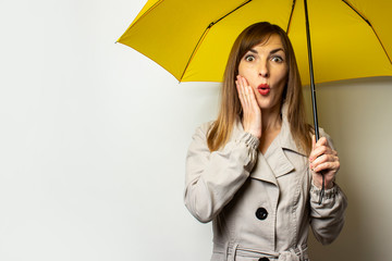 Portrait Young friendly woman in a classic cloak with a surprised face under a yellow umbrella on an isolated light background. Emotional face. Bad weather, rain, weather forecast