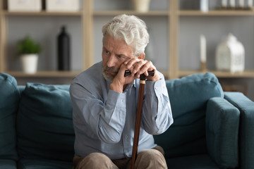 Tired upset mature man holding hands on walking stick, sitting on couch alone, exhausted unhappy older senior male using wooden cane during rehabilitation, elderly people healthcare - Powered by Adobe