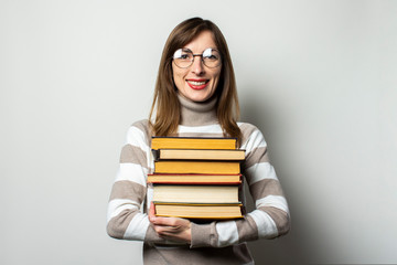 Portrait of a young friendly woman in a sweater and glasses holding a stack of books in her hands against an isolated light background. Emotional face. Education concept, exam preparation