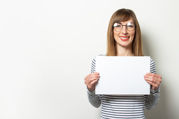 Naklejka premium Portrait of a young friendly woman in a long-sleeve sweater and glasses with a smile, holding a blank sheet of paper on an isolated light background. Emotional face