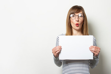 Fototapeta premium Portrait of a young friendly woman in a long-sleeve sweater and glasses with a surprised face holds a blank sheet of paper on light background. Emotional face. Gesture shock, surprise. Copy space