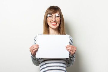 Naklejka premium Portrait of a young friendly woman in a long-sleeve sweater and glasses with a smile, holding a blank sheet of paper on an isolated light background. Emotional face