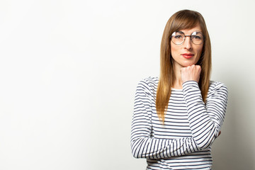 Obraz premium Portrait of a young friendly woman in a long sleeve sweater and glasses with a serious face holds her hand under her chin on an isolated light background. Emotional face. Gesture to think, dream