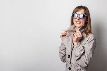 smiling young women in black sunglasses and wearing a beige cloak. Model holding on to the collar of a cloak and posing on a light background. Concept style, women's fashion. Empty space for text