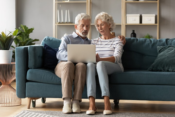 Mature man and woman using laptop together, looking at screen, older spouses browsing apps, shopping or chatting online, making video call, using bank service, sitting on cozy sofa in living room