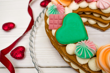 Festive dessert for birthday and Valentine's day.The cake in the shape of a heart decorated with meringue,gingerbreads and cream on the white wooden background with red hearts and ribbon