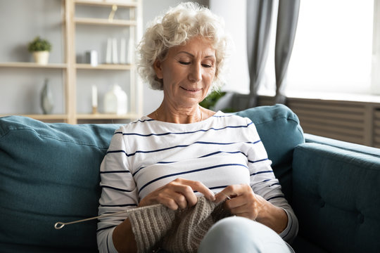 Satisfied Older Woman Knitting, Sitting On Cozy Sofa At Home, Enjoying Leisure Time On Weekend, Mature Senior Female Holding Knitting Needles, Creating Clothes, Elderly Generation Hobby Activity