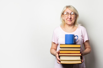 Portrait of an old friendly woman with a smile in a casual t-shirt and glasses holds a stack of books and a cup on an isolated light background. Emotional face. Concept book club, leisure, education