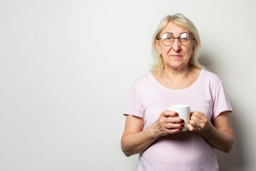 Portrait of an old friendly woman in casual t-shirt and glasses holding a mug in her hands on an isolated white background. Emotional face. Morning coffee concept, breakfast