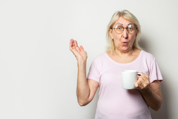 Old friendly woman with a surprised face in a casual t-shirt and glasses holding mug in his hands on isolated light background. Emotional face. Concept of morning coffee, breakfast, surprise, shock