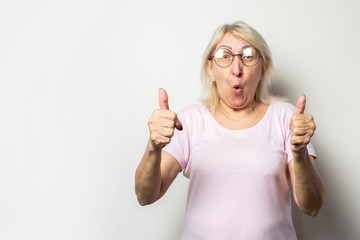 Portrait of an old friendly woman with a surprised face in a casual t-shirt and glasses makes a thumb up gesture on an isolated light background. Emotional face. Gesture is cool, everything is fine