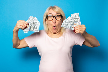 Portrait of an old friendly woman with a surprised face in a casual t-shirt and glasses holding money in her hands on an isolated blue background. Emotional face. Concept wealth, win, loan, pension