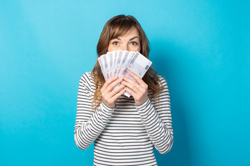 Portrait of a young friendly woman in casual t-shirt covered her face with a fan of money on an isolated blue background. Emotional face. Concept of wealth, win, credit. Gesture of joy from winning