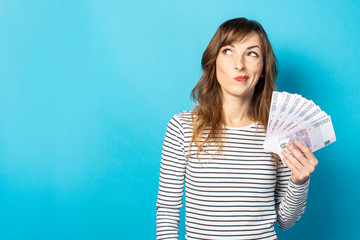 Young friendly woman in casual t-shirt holding money in her hands and looking to the side with an embarrassed face on isolated blue background. Concept of wealth, win, credit. Copy space