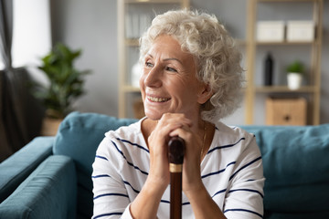 Fototapeta na wymiar Happy smiling older woman holding hands on wooden cane, looking into distance, in window, dreaming, excited mature female using walking stick, sitting on cozy sofa in living room, feeling positive