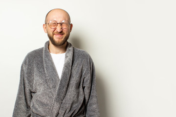 Portrait of a young bald man with a beard in a dressing gown and glasses closed his eyes, squinted at an isolated light background. Emotional face