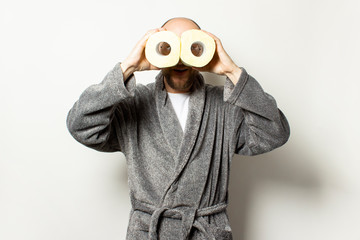 Portrait of young bald man with a beard in a dressing gown and glasses holds toilet paper in his hands like binoculars on light background. Emotional face. Bowel problems, constipation, hemorrhoids