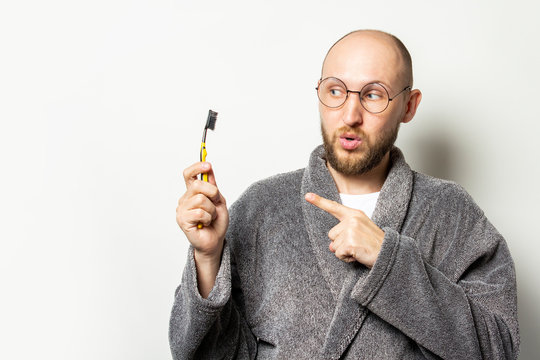 Portrait of young bald man with a beard in a dressing gown and glasses with a surprised face and points a finger at a toothbrush in his hand on light background. Oral care concept, personal hygiene