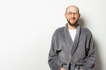 Portrait of a young bald man with a beard in a bathrobe and glasses on an isolated light background. Emotional face
