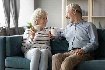 Happy mature couple drinking tea or coffee in cozy living room, holding cups, sitting on couch,...