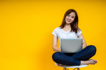 Young Asian woman teen smiling sitting on chair wearing t-shirt using laptop computer for work from home on covid-19 coronavirus crysis, studio shot on yellow background with copy space