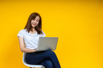 Fototapeta na wymiar Young Asian woman teen smiling sitting on chair wearing t-shirt using laptop computer for work from home on covid-19 coronavirus crysis, studio shot on yellow background with copy space