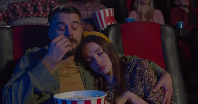 Close up view of pretty girl sleeping on her boyfriend shoulder during film in movie theatre. Man eating popcorn and embracing his asleep girlfriend while watching movie. Concept of leisure