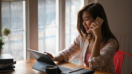 Beautiful woman talking on mobile while using a computer tablet and sitting at wooden working desk...
