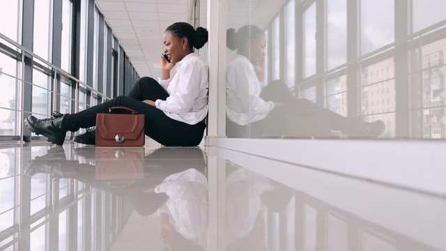 African-American woman is sitting on the floor in the airport.The young woman is waiting at the hall with a bag and chatting.