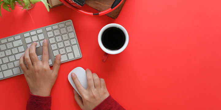 Top view image of hands typing on wireless keyboard and using a wireless mouse on red working desk surrounded by coffee cup, wireless headphone, old books and potted plant. Orderly workspace concept.
